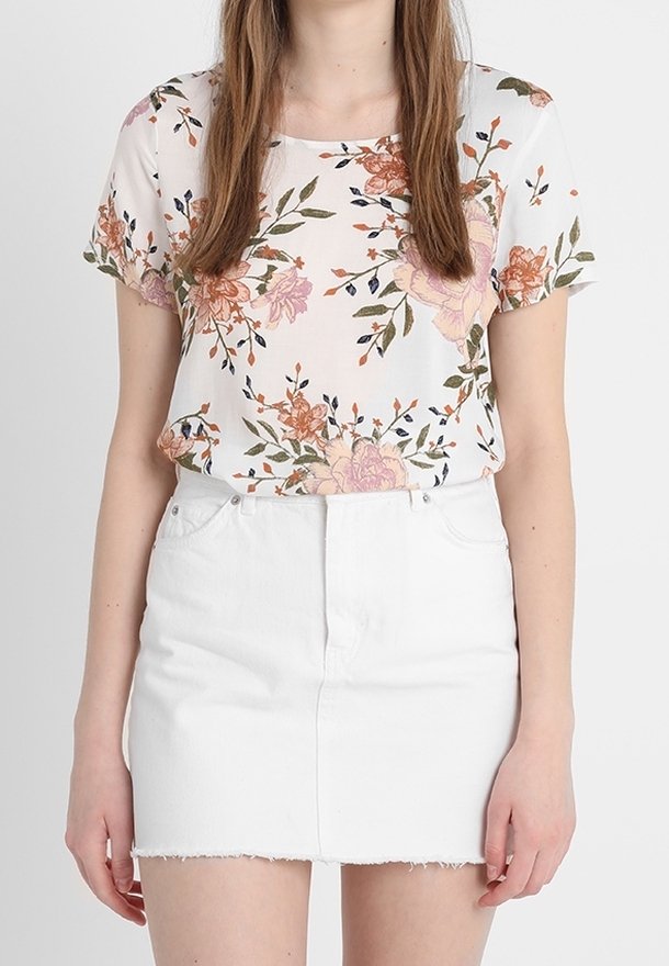 Mujer vestida con una blusa blanca de flores de manga corta metida dentro de una falda corta de mezclilla blanca con dobladillo desflecado, de pie contra un fondo liso.