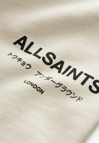 Beige stof med sort trykt tekst, "ALLSAINTS," med yderligere tekst på japansk og "LONDON" i bunden; glat tekstur.