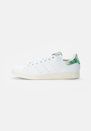 White low-top sneaker with perforated stripes, cream sole, and green patterned heel tab, viewed from the side on a plain background.