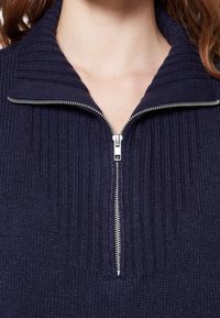 Maglione lavorato a maglia blu navy con colletto alto e zip frontale a metà. Presenta una texture a costine e una zip in metallo argentato.