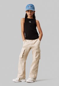 Top nero senza maniche con logo CK, pantaloni cargo beige con tasche, cappello di jeans azzurro chiaro e sneakers bianche. L'outfit è casual e funzionale.