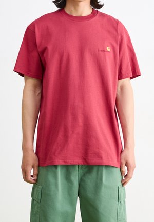 Person trägt ein locker sitzendes rotes Carhartt-T-Shirt und grüne Cargohosen, steht vor einem schlichten weißen Hintergrund.