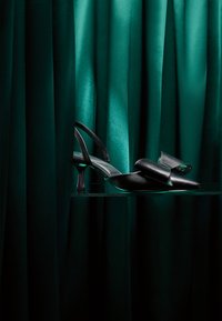 Black slingback high heel shoe with a large bow on the toe, displayed against dark green velvet curtains and a glass shelf.