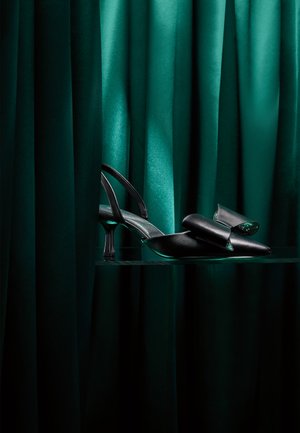 Mustat slingback-korkokengät, joissa on suuri bow-koriste varpaassa, esillä tummien vihreiden samettiverhojen ja lasihyllyn taustalla.