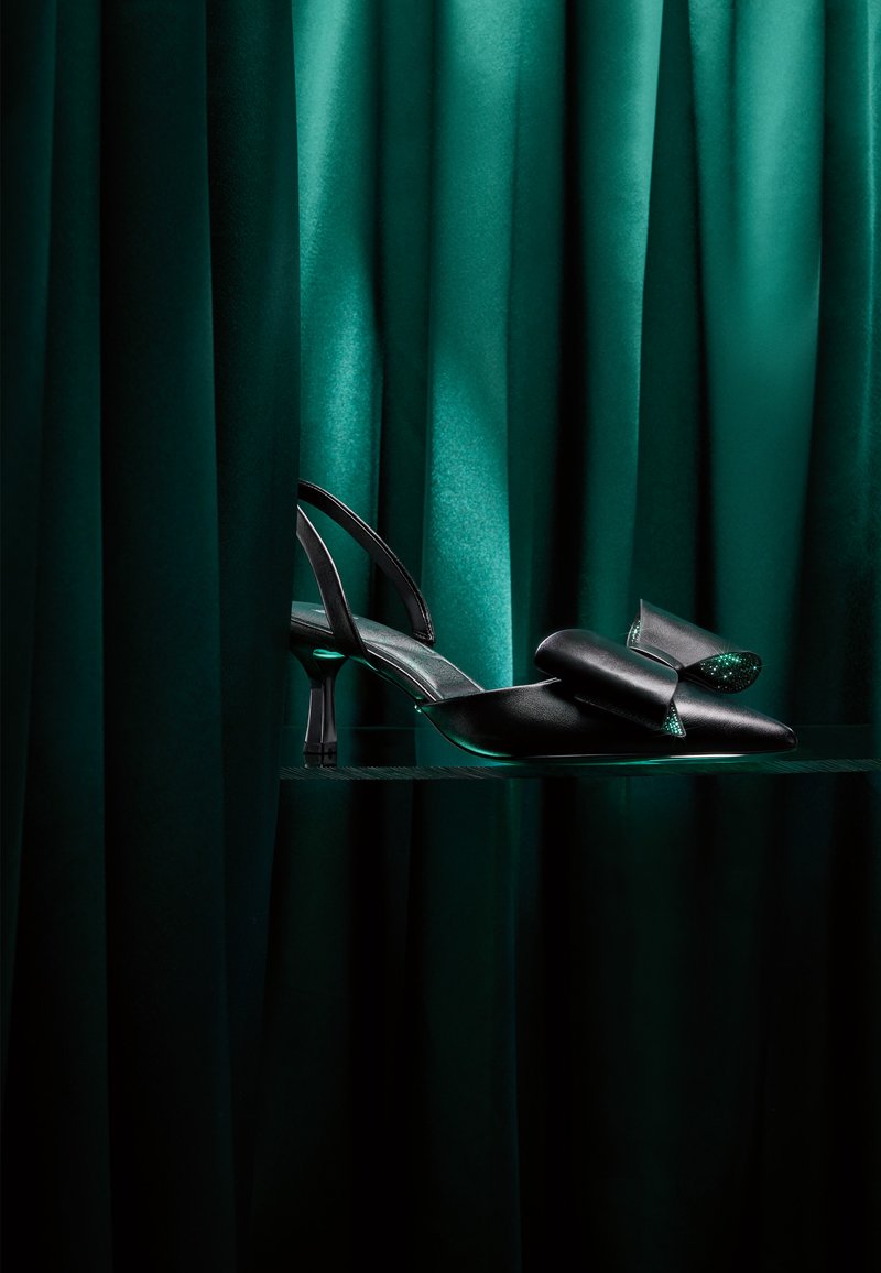 Black slingback high heel shoe with a large bow on the toe, displayed against dark green velvet curtains and a glass shelf.