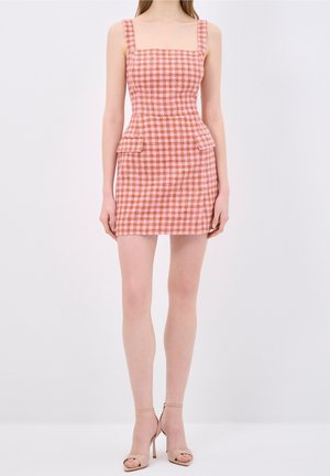 Kobieta ubrana w różowo-brązową, w kratę, bezrękawną mini sukienkę z przednimi kieszeniami i beżowe szpilki na pasku wokół kostki na białym tle.