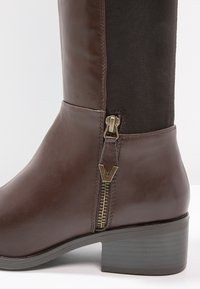 Botín de cuero marrón hasta el tobillo con parte trasera de textil, cuenta con un zipper dorado y un detalle de herrajes en forma de V. Tacón bajo y sólido. Acabado texturizado.