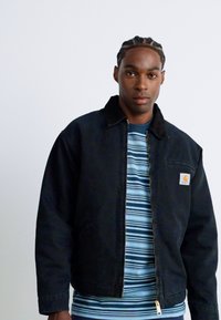 Junger Mann mit geflochtenem Haar, der eine schwarze Zip-Jacke über einem blau-weiß gestreiften Hemd trägt, steht vor einem einfarbigen hellen Hintergrund.