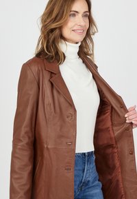 Bruine leren jas met een op maat gemaakt ontwerp, gekartelde revers en knoopsluiting. Gevoerde binnenkant en zakken, gedragen over een witte coltrui.