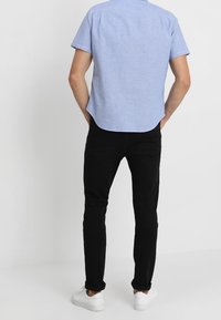 Camicia a maniche corte azzurro chiaro con orlo curvo, abbinata a jeans neri e sneakers bianche, vista da dietro.