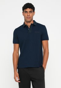 Camisa polo azul-marinho feita de tecido texturizado, com colarinho riscado a verde, placket de três botões e detalhe de logotipo bordado no peito.