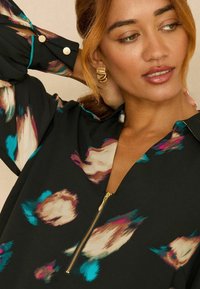 Fekete blúz színes, absztrakt mintával, rózsaszín, krémszín és türkiz árnyalatokban, cipzáros záródással és aranyszínű gombokkal az ujjakon.