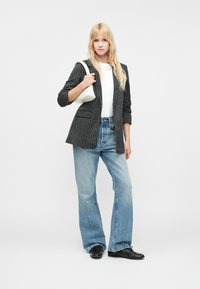 Fekete csíkos blazer, egy zsebbel, fehér póló, világoskék, bővülő farmernadrág és fekete cipő, egy krémszínű táskával a kezében.
