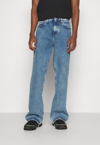 Blå denimjeans med en avslappnad, bred passform. Har en blekt tvätt, femfickorsdesign och en knappstängning. Svarta skor bärs under.