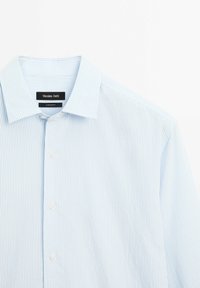 Chemise à fines rayures bleu clair et blanc avec boutons et col classique, présentée à plat sur une surface blanche.