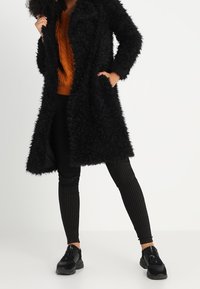 Sort lang frakke i falsk pels med blød tekstur, kombineret med en orange strikket sweater og sorte leggings med nålestriber samt sorte sneakers.