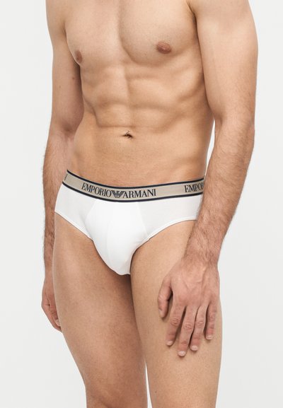 Ανδρικός κορμός που φορά άσπρα εσώρουχα Emporio Armani με μπεζ μέση και μαύρα γράμματα λογότυπου, όρθιος σε απλό φωτεινό φόντο.