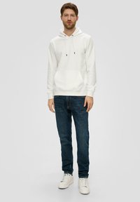 Haut en coton blanc avec poche kangourou, cordons de serrage ajustables, associé à un jean bleu foncé et des baskets blanches. Texture lisse, coupe décontractée.
