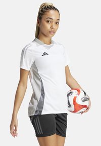 Maglietta sportiva bianca con dettagli grigi, maniche corte e collo tondo. Pantaloni corti neri e un pallone da calcio a motivi nella mano sinistra del modello.
