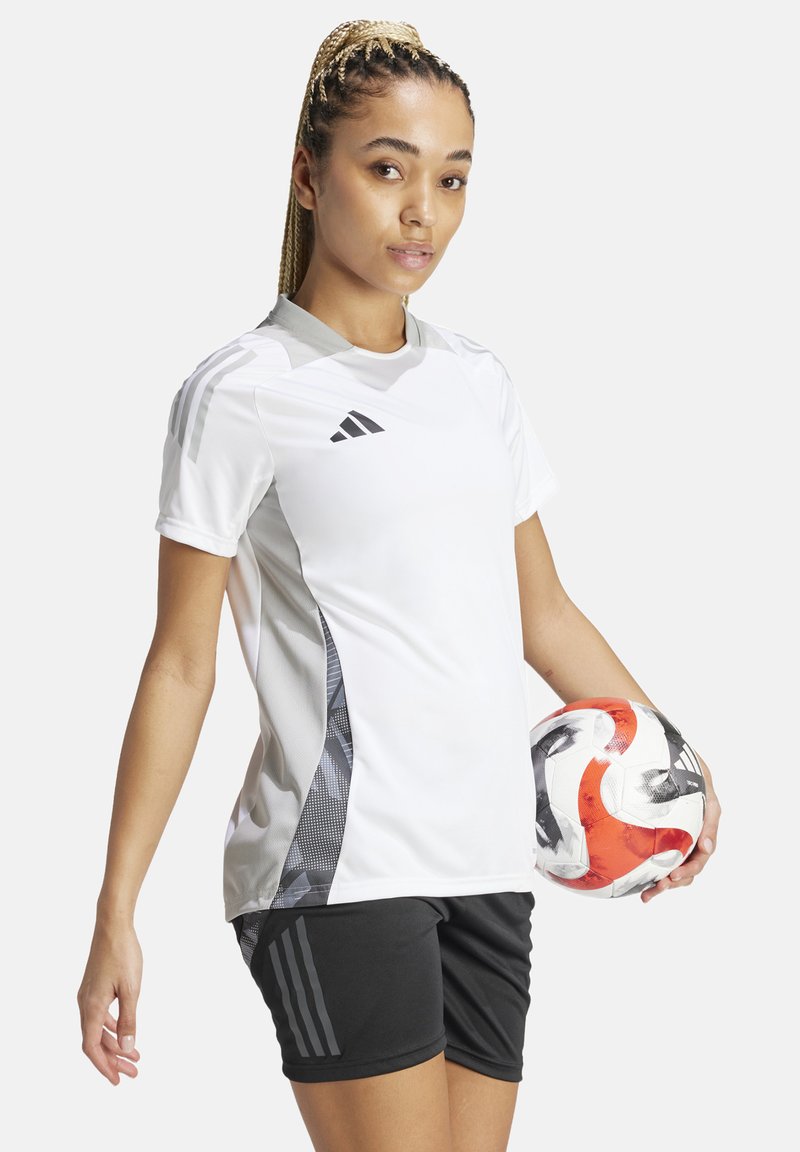 Maglietta sportiva bianca con dettagli grigi, maniche corte e collo tondo. Pantaloni corti neri e un pallone da calcio a motivi nella mano sinistra del modello.