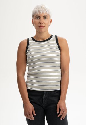 Persoon met kort wit haar die een mouwloos gestreept topje en zwarte broek draagt, staand tegen een effen witte achtergrond.