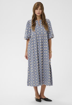 Žena stojí oblečená v modrých midi šatách s béžovým kvetinovým vzorom, nadýchanými lakťovými rukávmi, výstrihom s predným rozparkom, čiernymi špicatými balerínkami a zlatým náramkom.