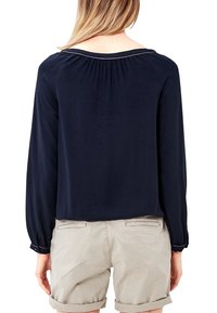 Blusa blu navy con maniche lunghe, tessuto testurizzato e scollo arricciato, abbinata a pantaloni shorts beige chiaro. La vista posteriore mostra il design e la vestibilità.