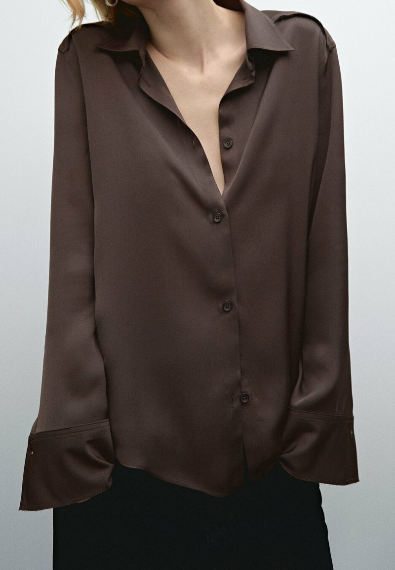 Femme portant un chemisier ample en satin brun foncé à boutons, avec de larges poignets et un col, visible du cou jusqu'aux hanches.