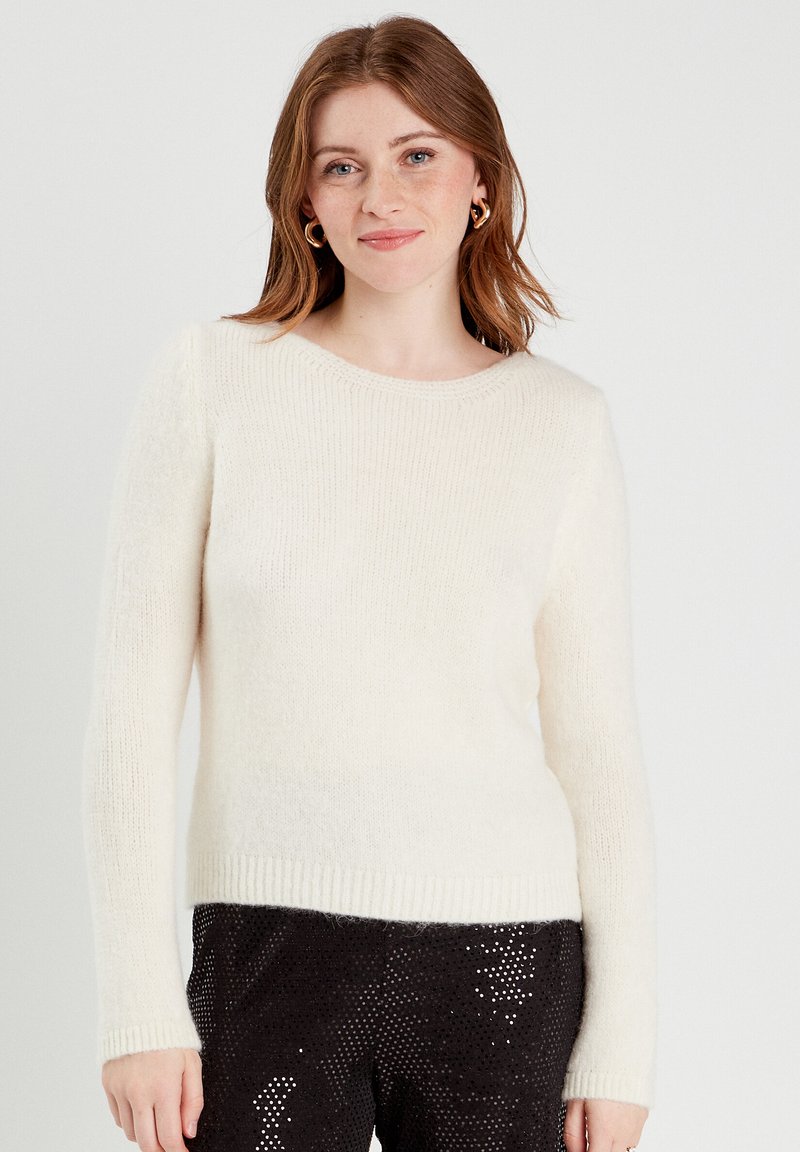Un pull crème en matériau doux et pelucheux, avec un col rond, des poignets et une ceinture côtelés ; associé à un pantalon noir pailleté.