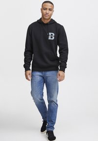 Sudadera negra con cordón y bolsillo canguro, con un audaz logo de la letra "B" en azul. Luciendo con jeans de color azul claro y zapatillas negras.