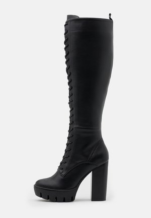 Botte en cuir noire montant jusqu'aux genoux avec un talon haut épais, un laçage frontal et une semelle épaisse crantée.