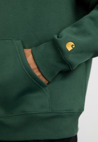 Zelena majica s rebrastim manžetama, koja na rukavu ima zlatni logotip. Materijal izgleda mekan i s teksturom, s džepom za kengure.
