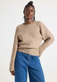 Pullover lavorato a maglia beige con un motivo testurizzato, scollo rotondo e maniche lunghe, abbinato a jeans a vita alta blu.