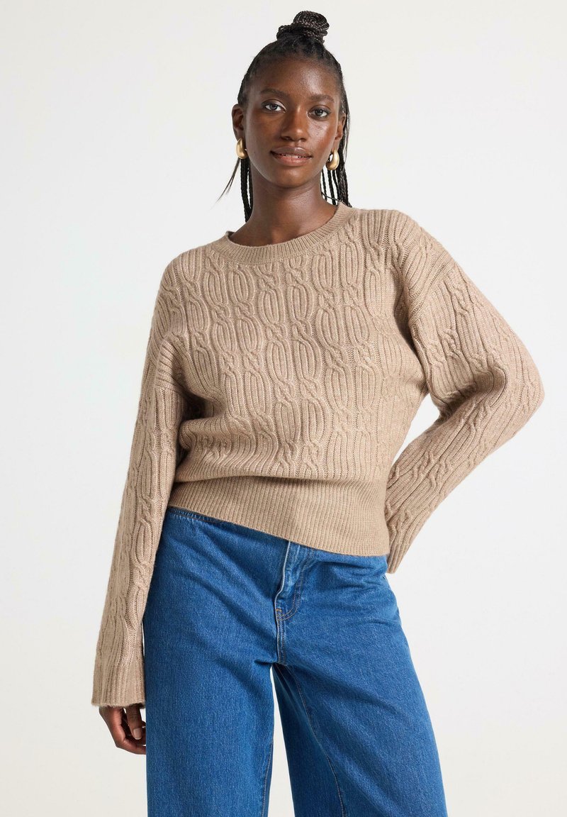 Pullover lavorato a maglia beige con un motivo testurizzato, scollo rotondo e maniche lunghe, abbinato a jeans a vita alta blu.