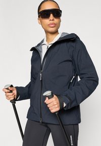 Tmavomodrá nepremokavá bunda so sivou kapucňou, predným zipsom a vreckami na zips. Čierne nohavice s bielym logom, drží turistické palice.