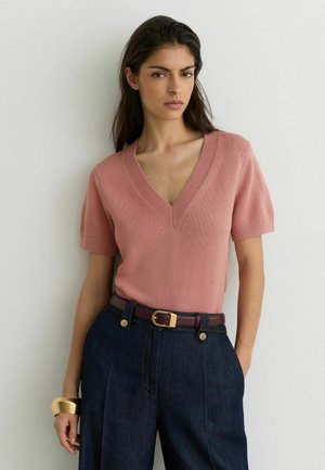 Naine, kellel on pikad tumedad juuksed, seljas roosa V-kaelusega lühikeste varrukatega kootud top, tumedad kõrge vöökohaga püksid pruuni rihmaga ja kuldne käevõru vasakul randmel.