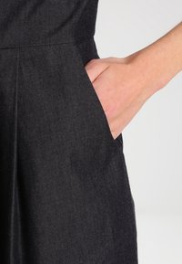 Musta farkkukangasmekko, jossa on sivutasku, pleatit ja sileä rakenne, korostaen sen jämäkkää muotoilua ja minimalistista tyyliä.