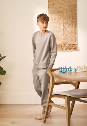 Băiat îmbrăcat în haine bej de relaxare, stând desculț lângă o masă din lemn cu piese de șah albastre și albe, într-o cameră luminoasă și minimalistă.