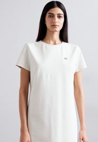 Biała bawełniana sukienka t-shirtowa z krótkimi rękawami, okrągłym dekoltem i małym logo na piersi. Gładka tekstura, luźny krój.