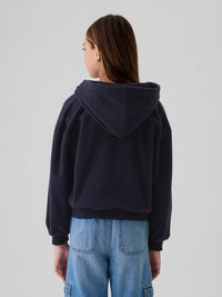 Jovem com cabelo castanho comprido, vestindo um hoodie azul marinho escuro e jeans de ganga azul claro, de costas contra um fundo cinza liso.