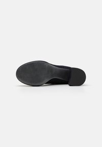 Chaussure à talon en suède noir avec une semelle lisse. Elle présente un bout arrondi et un large talon. Le dessous a un motif texturé pour une meilleure adhérence.