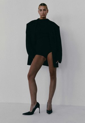 Kobieta stoi w stojącej pozycji przy gładkiej ścianie, ubrana w czarny, oversize'owy żakiet, czarny body i czarne szpilki, pewnie spoglądając przed siebie.