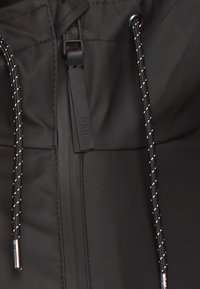 Svart vattentät jacka med en slät yta, utrustad med dragkedja och justerbara dragskon med reflekterande detaljer. Logotag synlig.