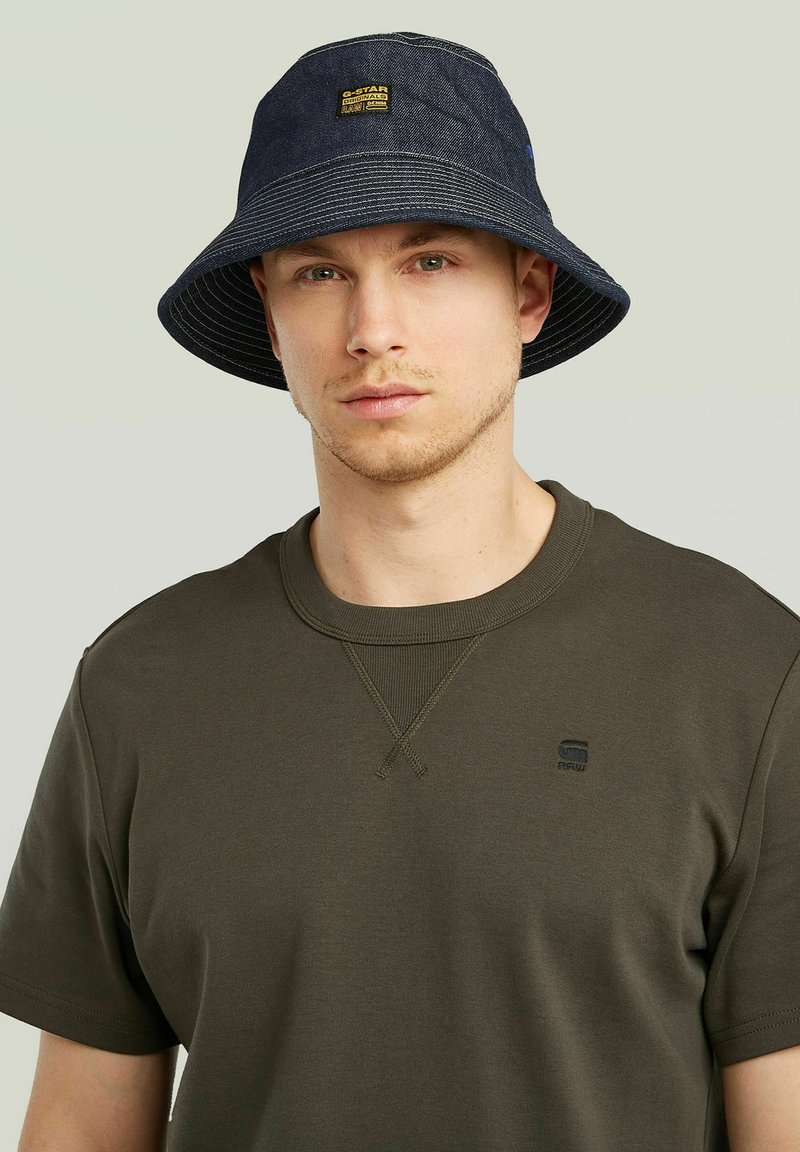 Bucket hat made of dark blue denim, featuring a wide brim and light striped interior. Logo patch on front. Worn with a dark green shirt.