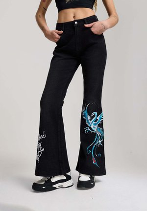 Person wearing black flared jeans with blue bird embroidery and white script on legs, paired with black and white sneakers.