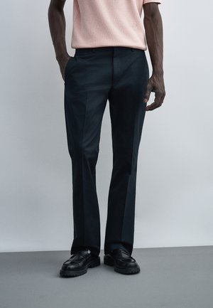 Mand iført sorte klassiske bukser, sorte læder loafers og en lyserød kortærmet skjorte stoppet ind i bukserne med hænderne i lommerne.