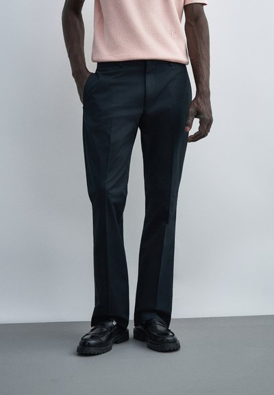 Homme portant un pantalon de costume noir, des mocassins en cuir noirs et une chemise rose clair à manches courtes rentrée dans le pantalon, les mains dans les poches.