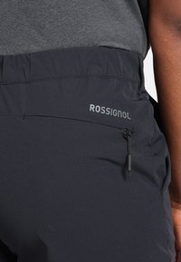 Pantalons de plein air noirs avec une texture lisse, une taille élastique et une poche zippée sur le côté. La marque "ROSSIGNOL" est imprimée en argent.