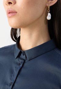 Camicia blu navy con colletto e chiusura a bottoni, abbinata a un orecchino ad anello dorato con una perla a forma di goccia testurizzata.