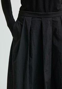 Osoba w czarnej bluzce z długim rękawem i wysokich, czarnych plisowanych spódnicy, z rękami w kieszeniach, pokazana od talii do połowy uda.